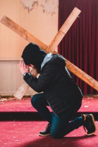 A young man is wearing a black coat, hat, and jeans and kneels in a ruined church while carrying a cross on his back. The reality is that all instances of running, staying, and returning are ultimately in God's hands…It is crucial to understand God's will, have a clear heavenly vision, and discern the mysteries behind our circumstances…Whether we choose running, staying, or returning, we should choose the path of death on the cross.